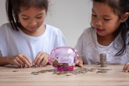Two cute asian child girls putting money into piggy bank to save money for the future together with fun and happinessの写真素材