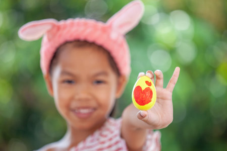 Happy asian child girl drawing heart picture on egg and holding painted easter egg with fun in...