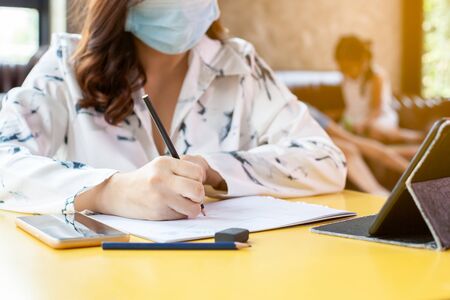 Work from home.Woman in quarantine for Coronavirus COVID-19 wearing protective mask writing on notebooks working at home while her kids playing at home during coronavirus outbreakの写真素材