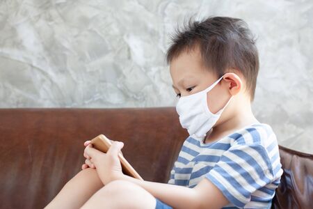 Asian child boy wearing protection mask plays on the smartphone at home quarantine from the coronavirus Covid-19 and air pollution pm2.5.の写真素材