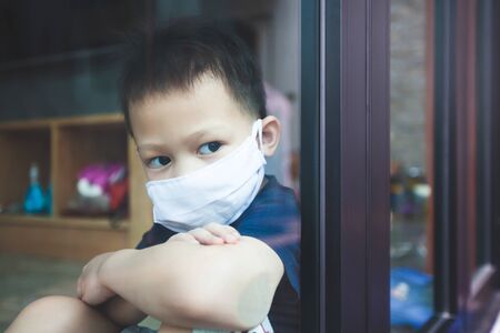 Asian child boy wearing protection mask looking outside through the window and stay at home quarantine from the coronavirus Covid-19 and air pollution pm2.5.の写真素材