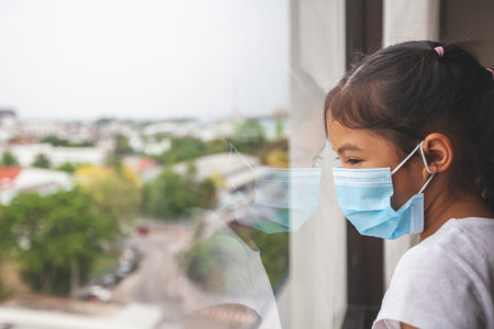 Asian child girl wearing protection mask looking outside through the window and stay at home quarantine from the coronavirus Covid-19 and air pollution pm2.5.の写真素材