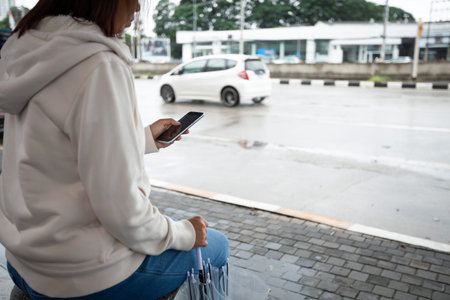 Asian woman is using on smartphone, checking social media network  while waiting taxi at bus stop in...