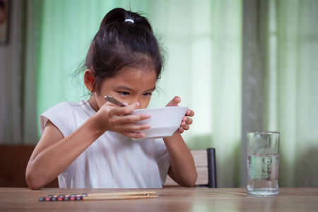 Cute asian child girl eating delicious instant noodles and drinking soup from bowl for her lunch in the houseの写真素材