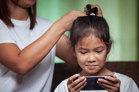 Asian child girl playing on her smartphone while mother is making hair on her head to get ready for school.の写真素材