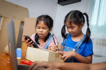 Woman hand using notebook computer taking receive and checking online purchase shopping order. Child is writing address into the box to help parent prepares delivery box for the online customer.の写真素材