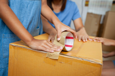 Close up on child hand helping her parent to pack the stuffs and tape a box before moving to new...