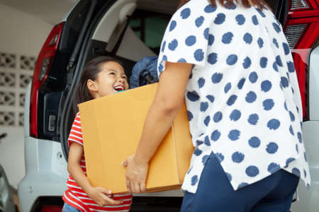 Happy asian child girl and mother helping to carry a cardboard box with stuffs moving into the car together to relocation on moving day. Home renovation and relocation concept.の写真素材