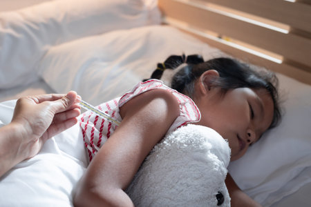 Sick asian child girl is sleeping on bed and her mother is checking her body temperature with...