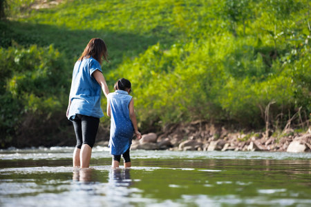 Asian mother holding her child hand to walk and play in the river together with fun and enjoy with...