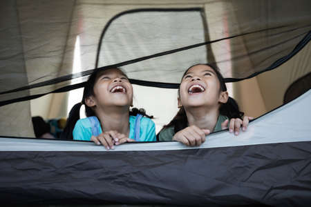 Two cute asian child girl lying down and playing together in tent on campsite while camping with family. Holiday and travel family camping concept.の写真素材