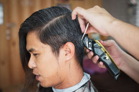 Asian young man being cut from long hair to short hair with electric clipper machine by professional barber in barbershop.の写真素材