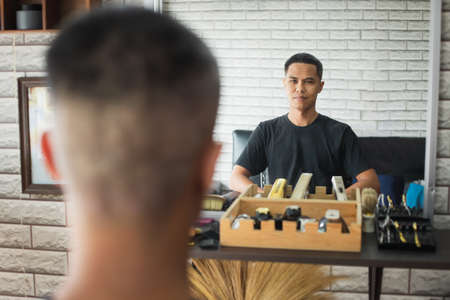 Asian young man sitting on chair and looking himself in mirror after finished haircut in barbershop.の写真素材