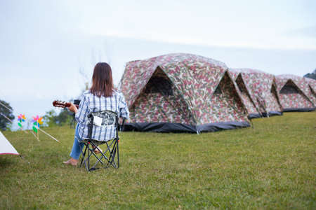 Asian woman sitting on picnic chair and playing guitar while camping with family in the camping site in the beautiful nature. Family enjoy with nature and camping concept.の写真素材