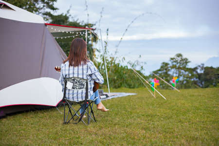 Asian woman sitting on picnic chair and playing guitar while camping with family in the camping site in the beautiful nature. Family enjoy with nature and camping concept.の写真素材