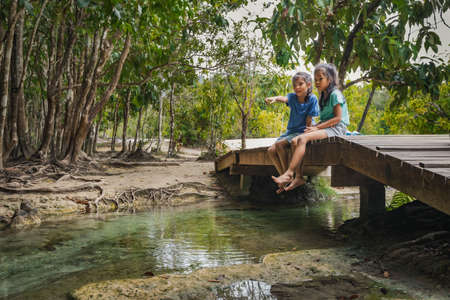 Two cute asian child girls sitting on the wooden bridge over the river and enjoy with nature together in summer time.の写真素材