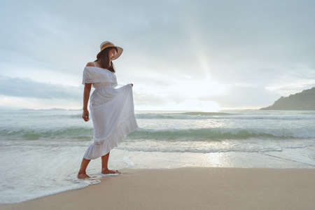 Asian woman wearing stylish hat and clothes walking on the beach and enjoying beautiful nature in the sunset time. Summer holidays and travel concept.の写真素材