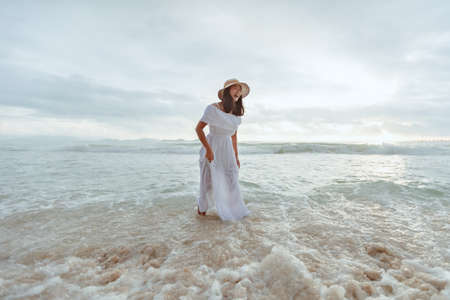 Asian woman wearing stylish hat and clothes laughing while playing water in the sea with happiness and enjoying beautiful nature in the sunset time. Summer holidays and travel concept.の写真素材