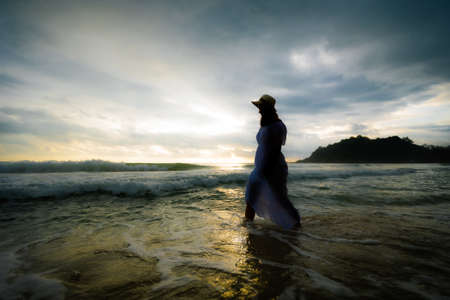 Silhouette of asian woman wearing stylish hat and clothes walking on the beach and enjoying beautiful nature in the sunset time. Summer holidays and travel concept.の写真素材