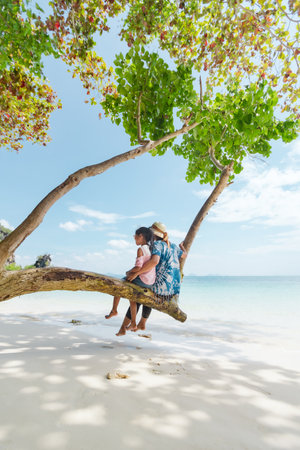 Asian mother and child girl sitting on tree and enjoying with beautiful sea nature together in their vacation. Summer holidays and family travel concept. Travelling in Thailand.の写真素材