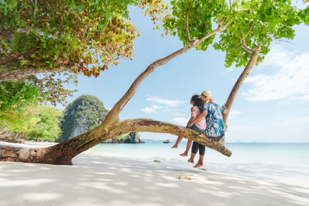 Asian mother and child girl sitting on tree and enjoying with beautiful sea nature together in their vacation. Summer holidays and family travel concept. Travelling in Thailand.の写真素材
