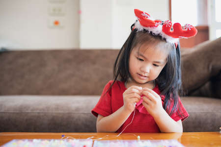 Cute asian child girl wearing christmas costume threading beads onto a string with intention and fun in home.の写真素材