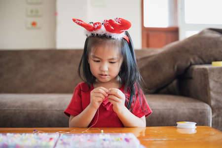 Cute asian child girl wearing christmas costume threading beads onto a string with intention and fun in home.の写真素材