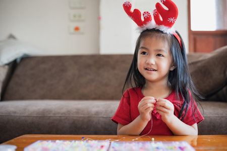 Cute asian child girl wearing christmas costume threading beads onto a string with intention and fun in home.の写真素材
