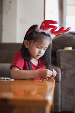 Cute asian child girl wearing christmas costume threading beads onto a string with intention and fun in home.の写真素材