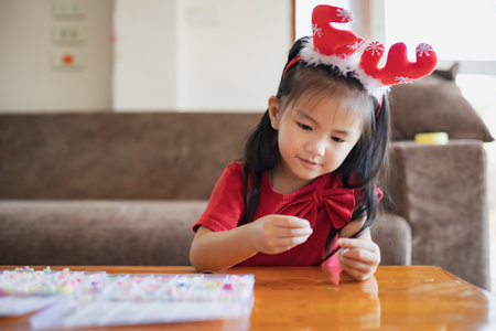 Cute asian child girl wearing christmas costume threading beads onto a string with intention and fun in home.の写真素材