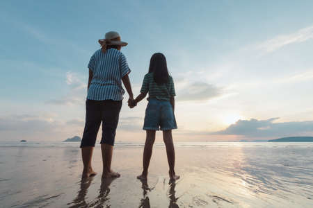 Silhouette of asian mother and daughter holding hand together standing on the beach looking at the beautiful sea and sky at the sunset time.の写真素材