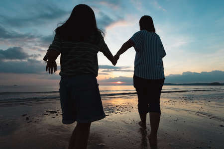 Silhouette of asian mother and daughter holding hand and walking on the beach together at the sunset time with beautiful sea and sky. Family enjoy with nature concept.の写真素材