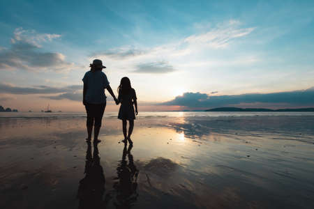 Silhouette of asian mother and daughter holding hand and walking on the beach together at the sunset time with beautiful sea and sky. Family enjoy with nature concept.の写真素材