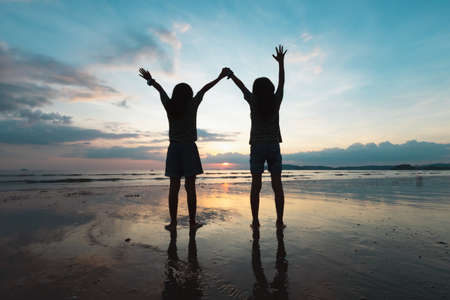 Silhouette of two asian child girls playing on the beach together at the sunset time with beautiful sea and sky. Family enjoy with nature concept.の写真素材