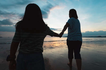 Silhouette of asian mother and daughter holding hand and walking on the beach together at the sunset time with beautiful sea and sky. Family enjoy with nature concept.の写真素材