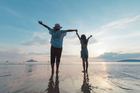 Silhouette of asian mother and daughter holding hand together standing on the beach looking at the beautiful sea and sky at the sunset time.の写真素材