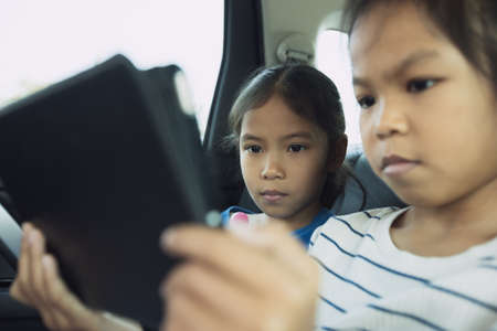 Two asian child girls sitting on backseat and watching cartoon in tablet together while traveling in the car with fun. Childhood and family lifestyles in car concept.の写真素材