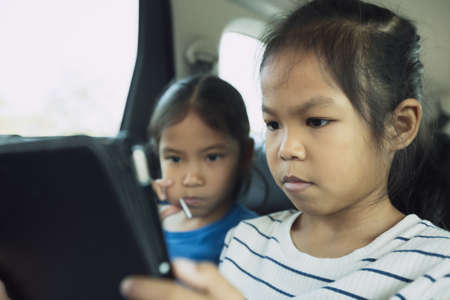 Two asian child girls sitting on backseat and watching cartoon in tablet together while traveling in the car with fun. Childhood and family lifestyles in car concept.の写真素材