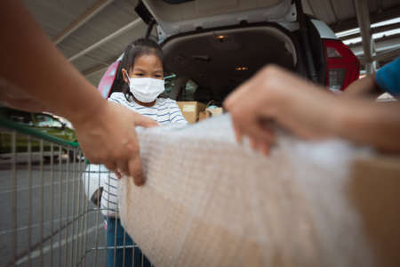 Asian mother and two daughters wearing protection mask helping to put stuffs in the shopping trolley into trunk of car together in parking during coronavirus pandemic as new normal lifestyle.の写真素材