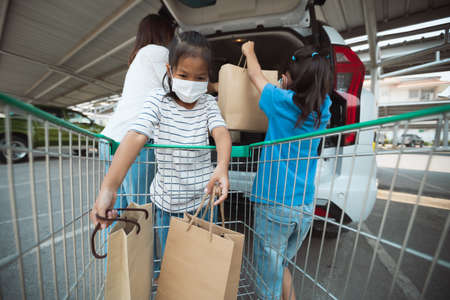 Asian mother and two daughters wearing protection mask helping to put stuffs in the shopping trolley into trunk of car together in parking during coronavirus pandemic as new normal lifestyle.の写真素材