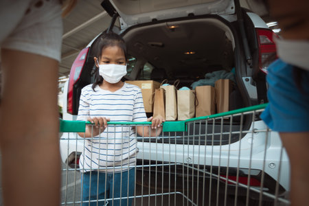 Asian mother and two daughters wearing protection mask helping to put stuffs in the shopping trolley into trunk of car together in parking during coronavirus pandemic as new normal lifestyle.の写真素材