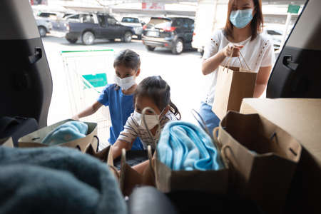 Asian mother and two daughters wearing protection mask helping to put stuffs in the shopping trolley into trunk of car together in parking during coronavirus pandemic as new normal lifestyle.の写真素材