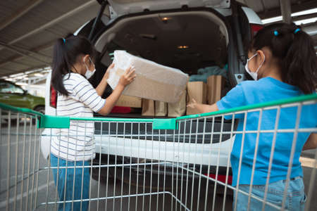 Asian mother and two daughters wearing protection mask helping to put stuffs in the shopping trolley into trunk of car together in parking during coronavirus pandemic as new normal lifestyle.の写真素材