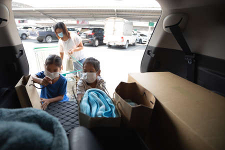 Asian mother and two daughters wearing protection mask helping to put stuffs in the shopping trolley into trunk of car together in parking during coronavirus pandemic as new normal lifestyle.の写真素材