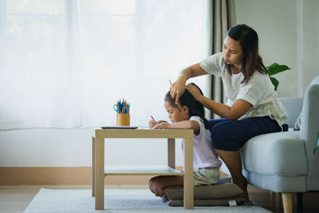 Asian child girl doing homework and mother is combing her hair in the living room.の写真素材