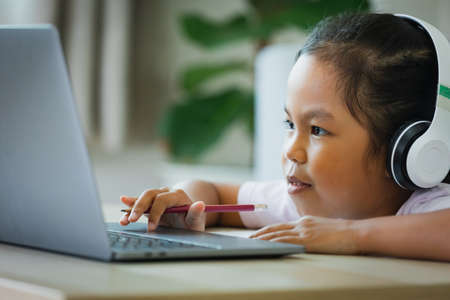 Asian child girl student wear headphone study online with teacher by video call. Child is homeschooling with computer laptop during quarantine due to pandemic.の写真素材