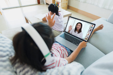 Asian kid sitting on sofa and having video conference chat with teacher and class group with sister...