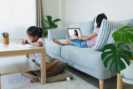 Asian kid sitting on sofa and having video conference chat with teacher and class group with sister doing worksheet stay beside. Children study from home during quarantine due to virus outbreak.の写真素材
