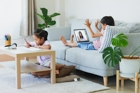 Asian kid sitting on sofa and having video conference chat with teacher and class group with sister doing worksheet stay beside. Children study from home during quarantine due to virus  outbreak.の写真素材