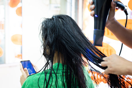 Young asian woman wearing protection mask and using smartphone while she getting a drying hair with hair dryer by hairdresser. Hairdresser drying hair to client at hair salon.の写真素材